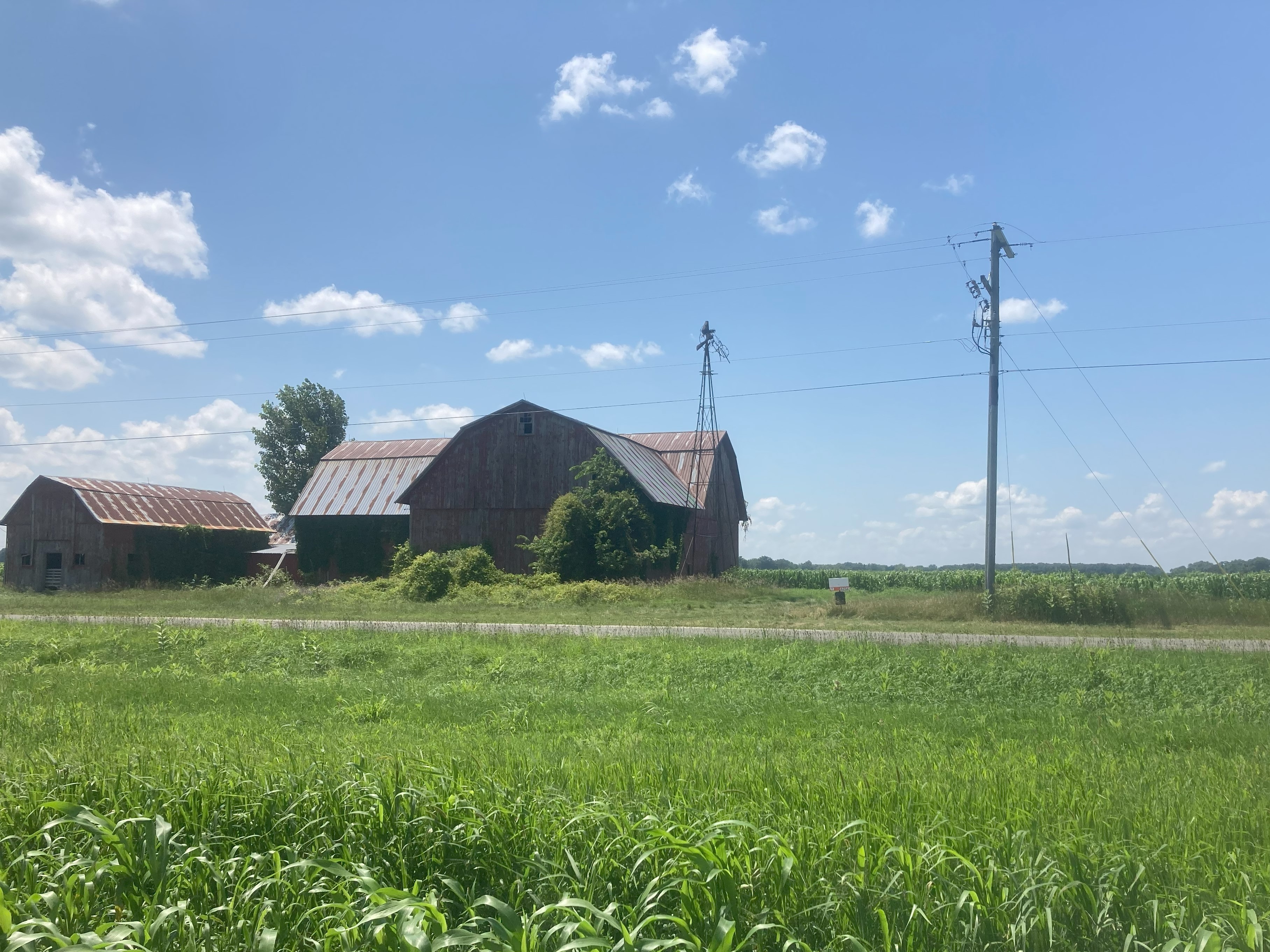 An older red barn and adjacent outbuilding sit beside a rural road, surrounded by grass and fields under a bright blue sky with scattered clouds.